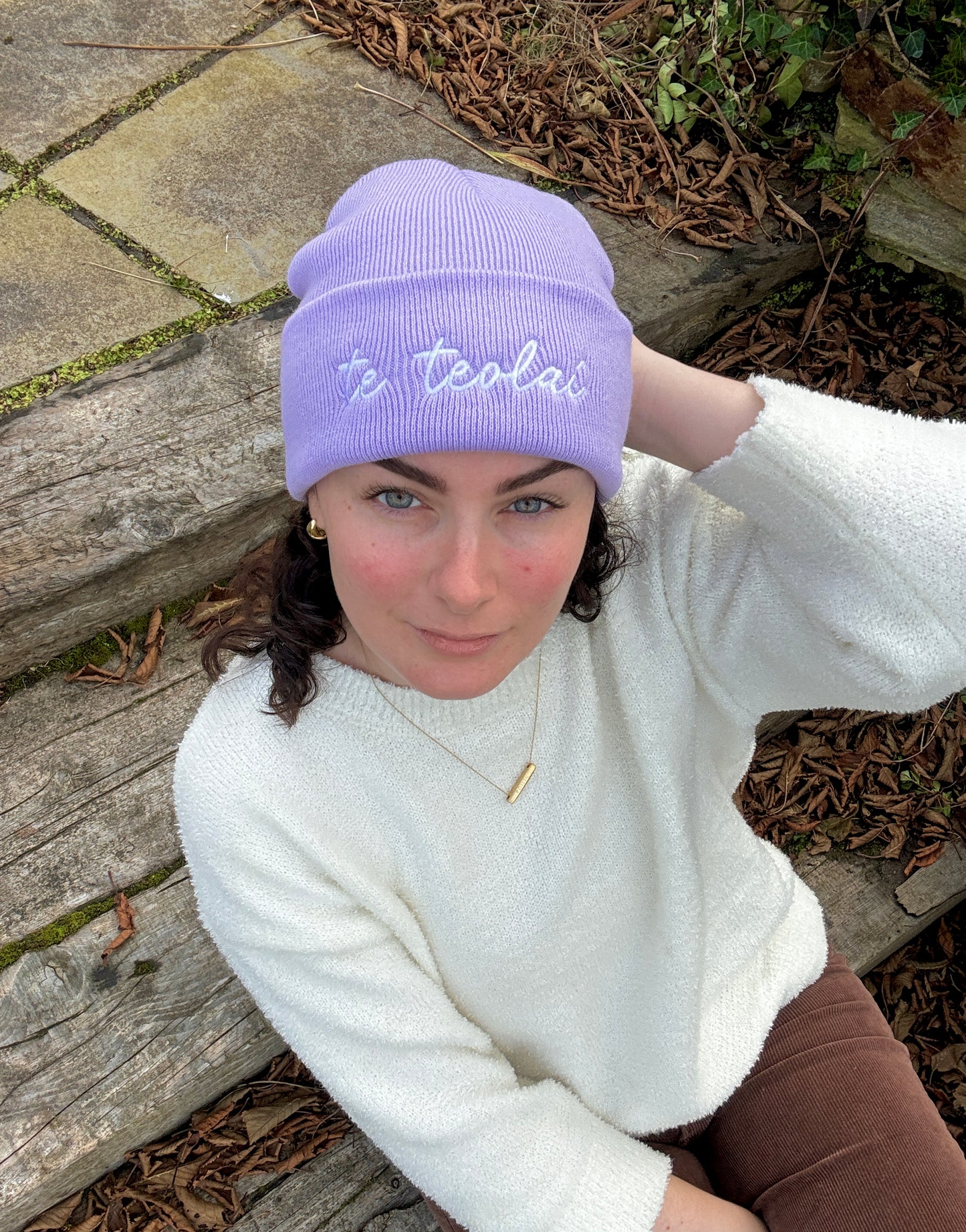 Person wearing a purple beanie with 'te teolaí' text, sitting outdoors on a stone surface.