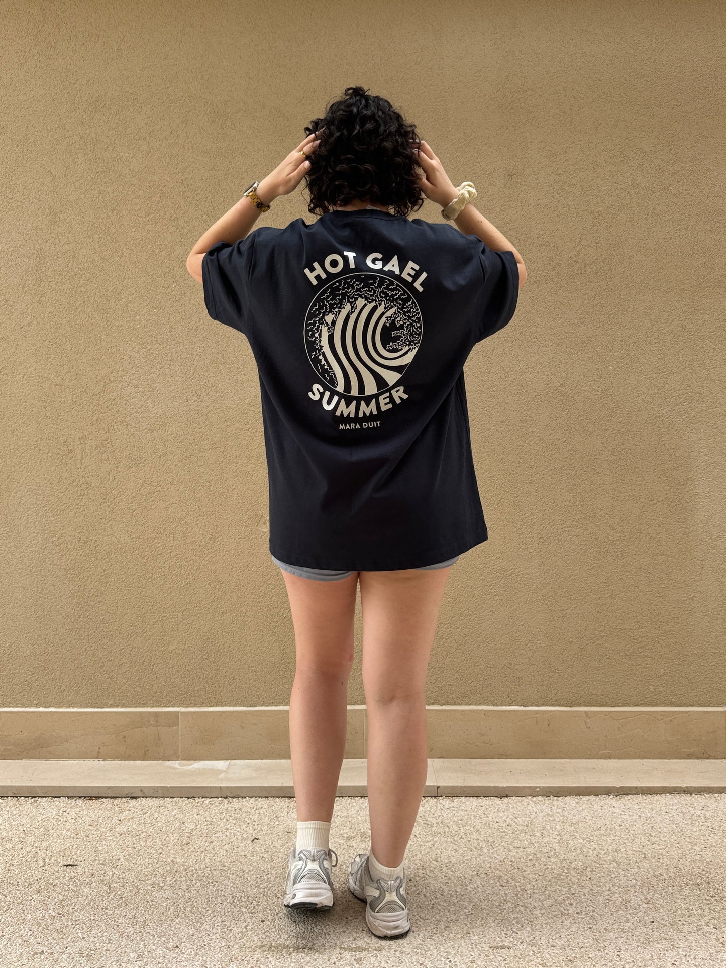 Person wearing a black Hot Gael Summer t-shirt with text and graphics on the back against a beige wall.