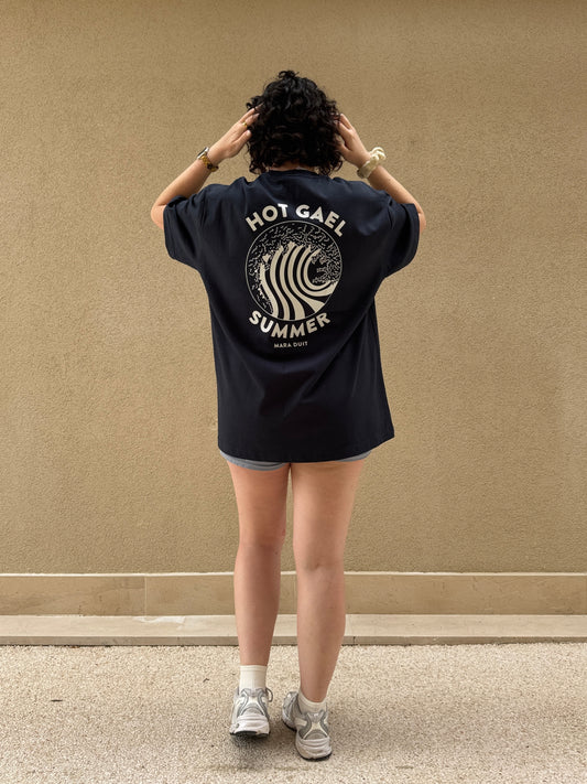 Person wearing a black Hot Gael Summer t-shirt with text and graphics on the back against a beige wall.
