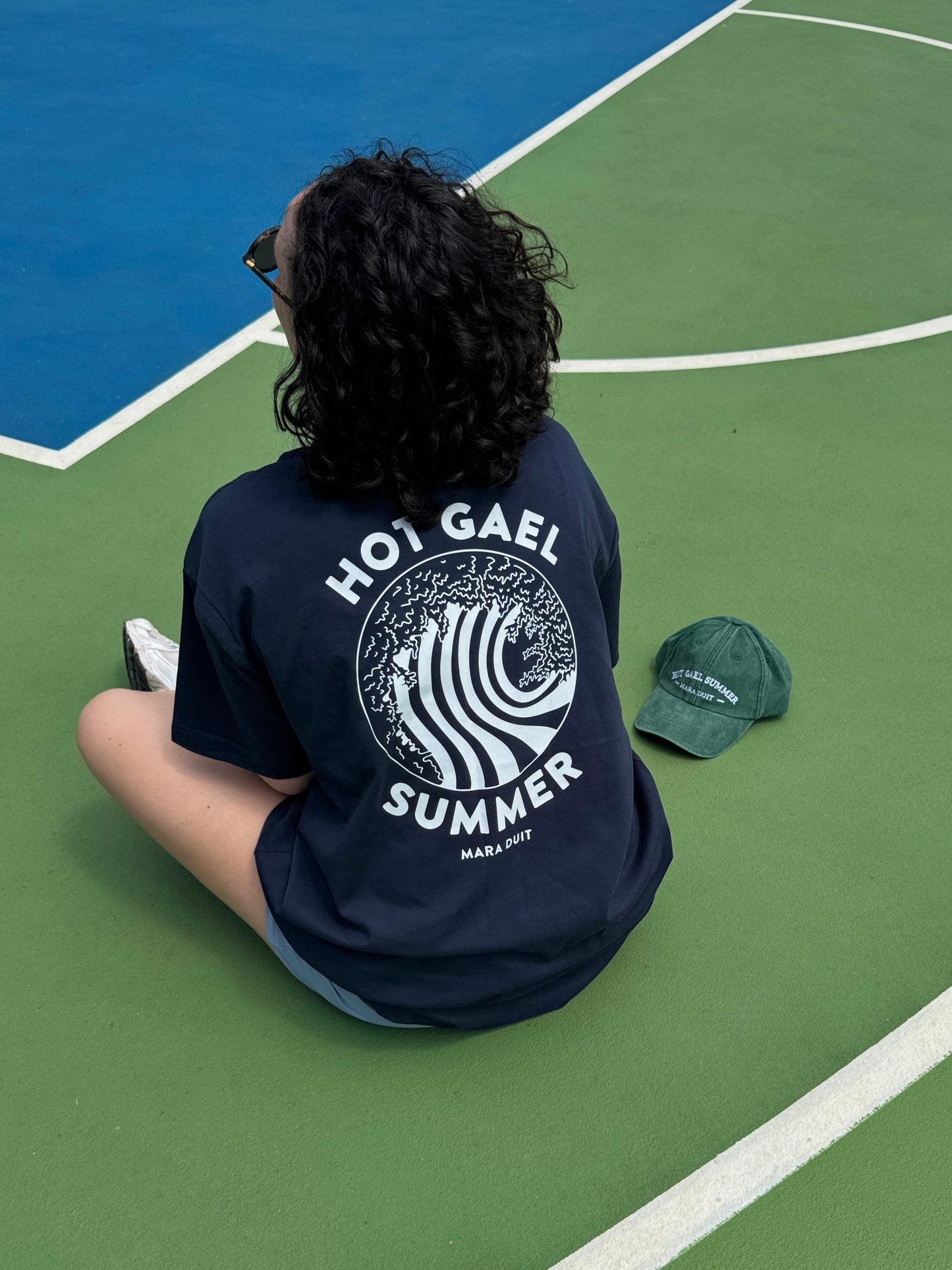 Person wearing a black t-shirt with 'Hot Gael Summer' text and wave design on a green basketball court.