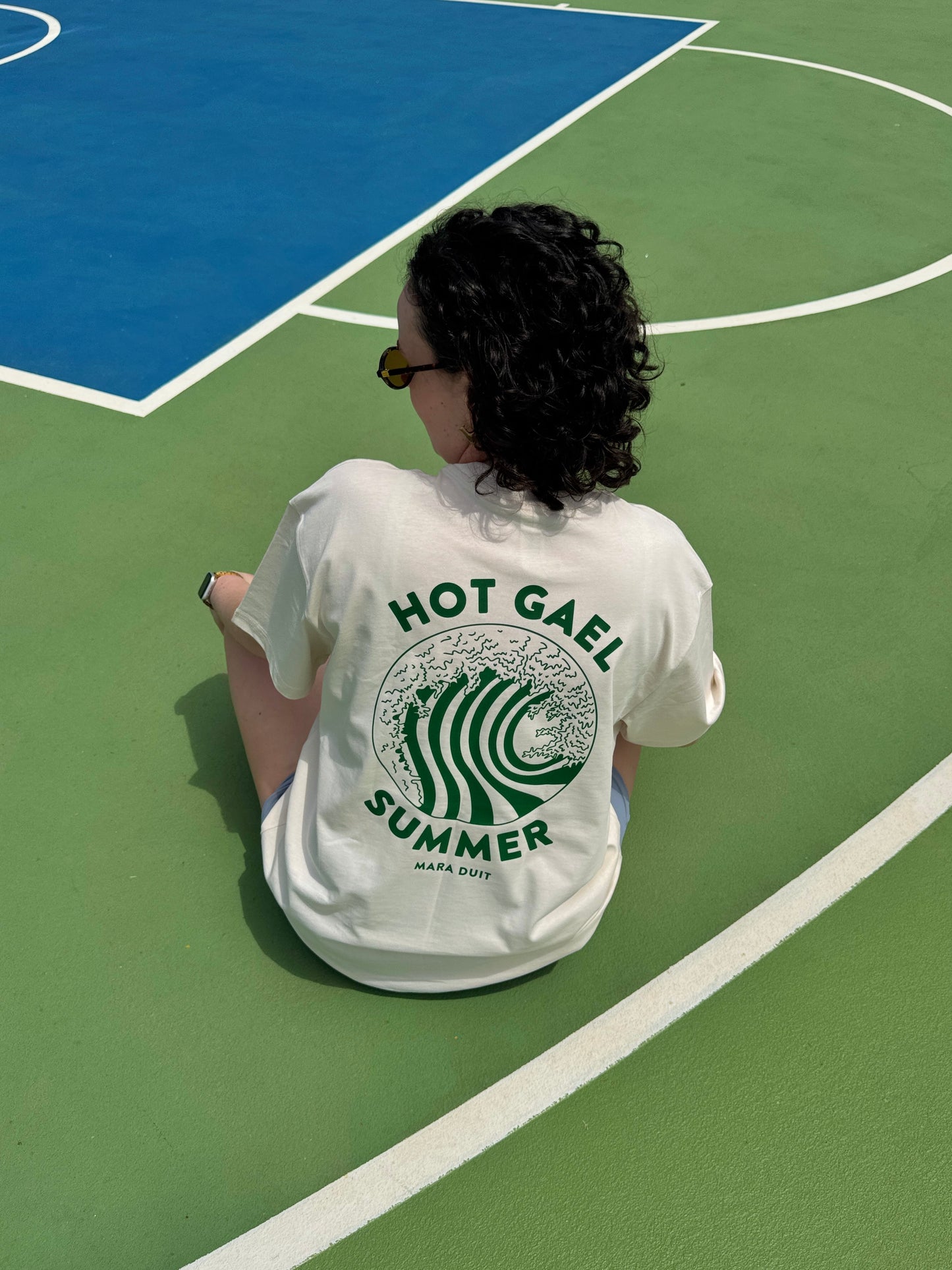 Person wearing a cream t-shirt with 'Hot Gael Summer' design on a green basketball court.