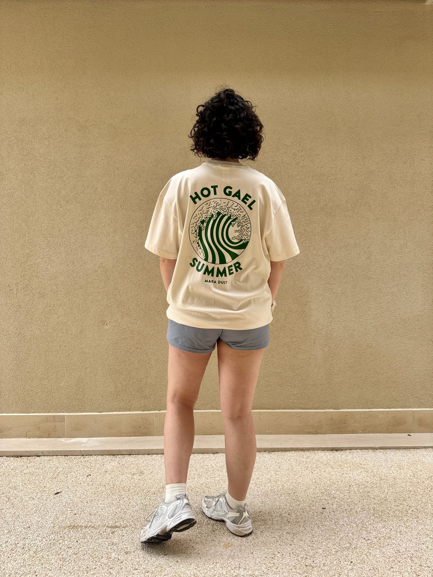 Person wearing a cream t-shirt with hot gael summer text and graphics, standing against a plain wall.