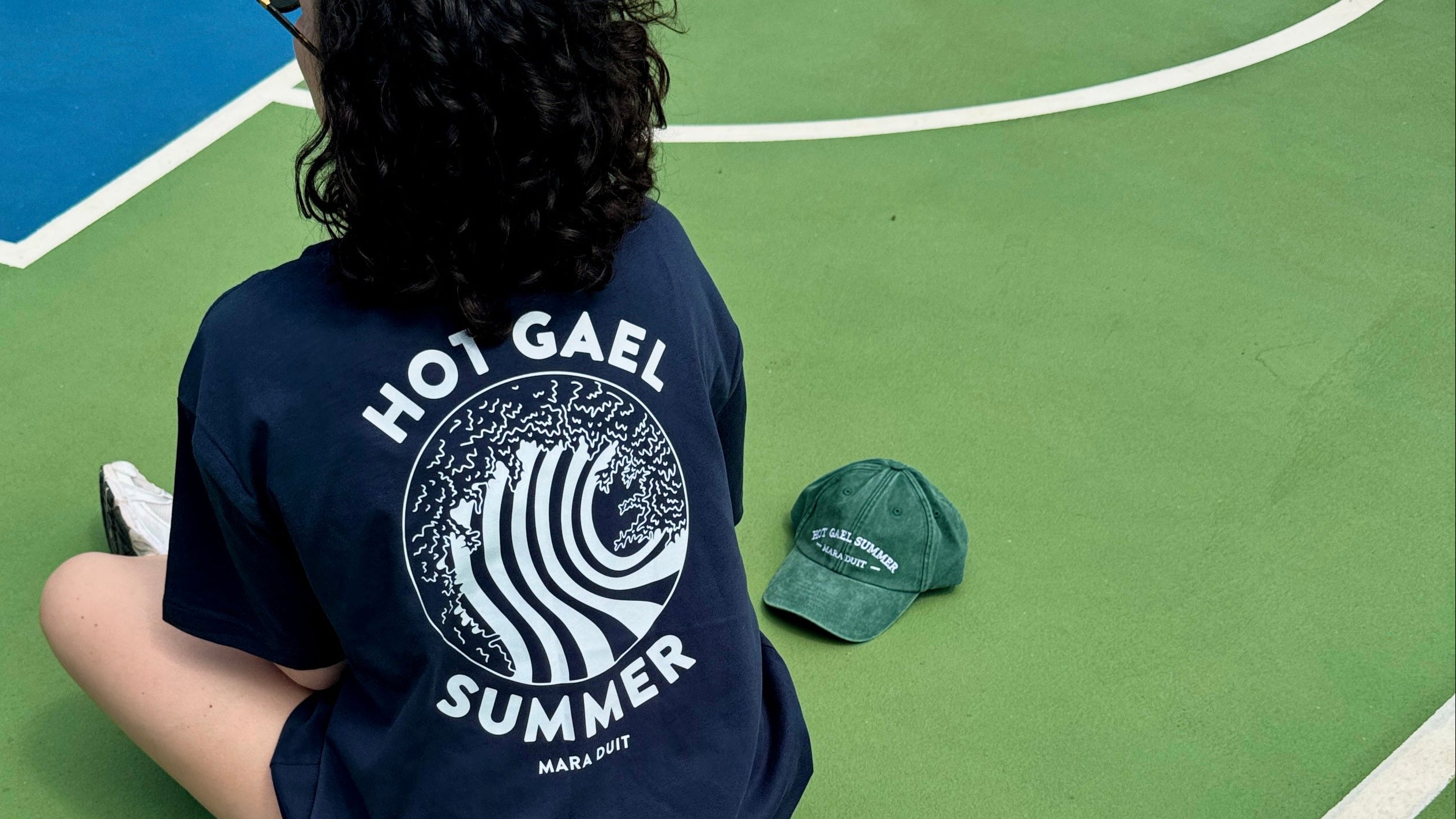 Person wearing a navy blue t-shirt with 'Hot Gael Summer' design, sitting on a basketball court.