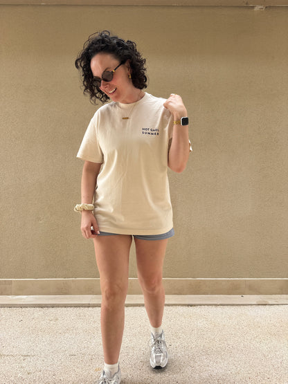 Person wearing a beige Hot Gael Summer t-shirt and shorts standing against a plain wall.