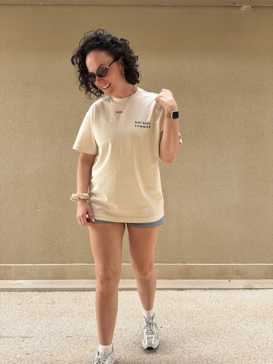 Person wearing a beige Hot Gael Summer t-shirt and shorts standing against a plain wall.