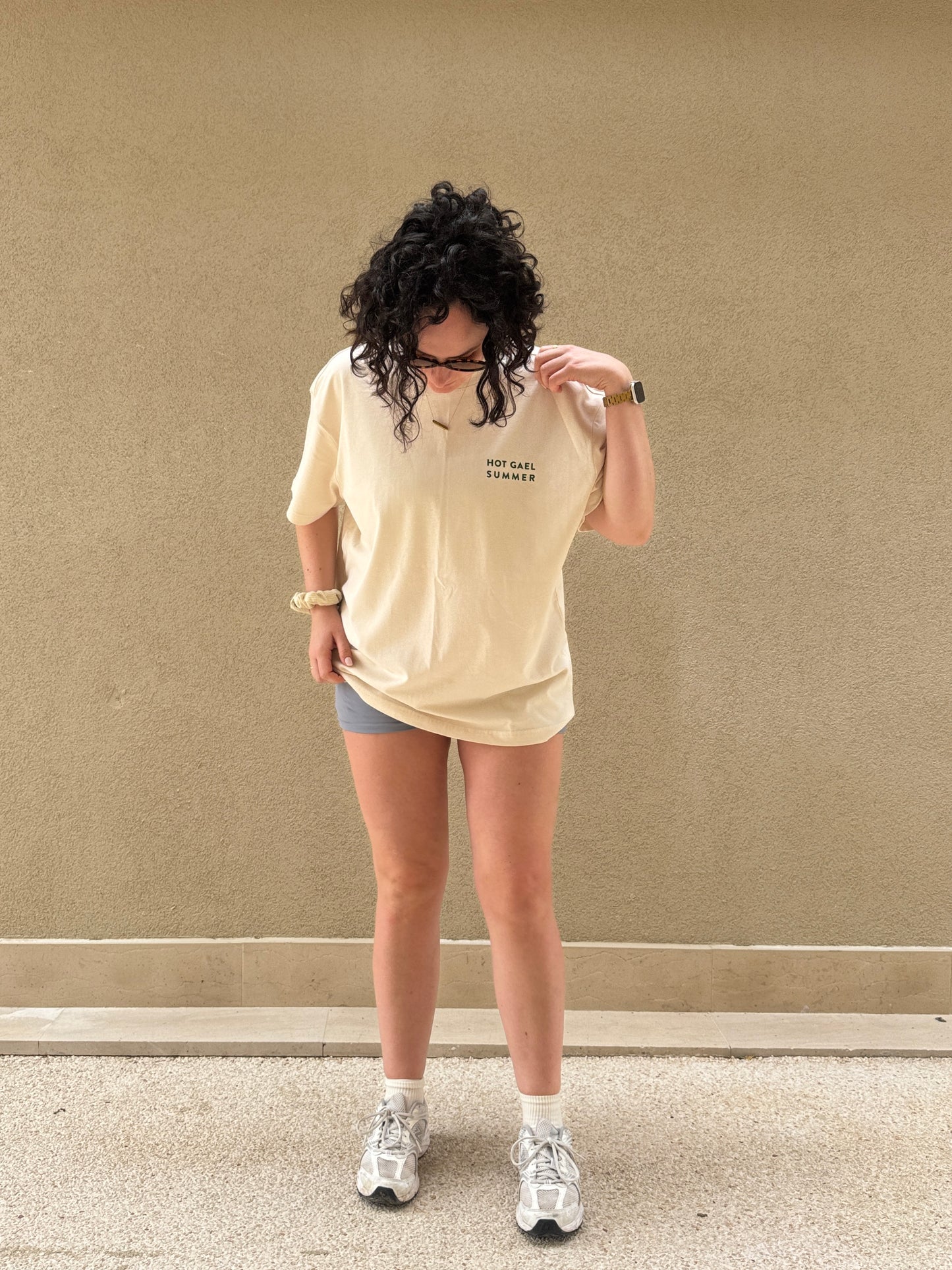 Person wearing a cream t-shirt with hot gael summer text, shorts, and white sneakers against a beige wall.