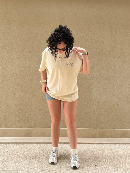 Person wearing a cream t-shirt with hot gael summer text, shorts, and white sneakers against a beige wall.