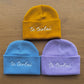 Three beanies in yellow, blue, and purple with 'te teolaí' embroidery on a wooden surface.