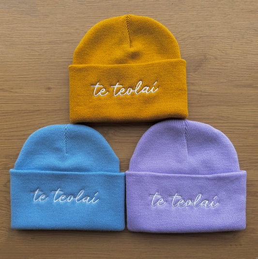 Three beanies in yellow, blue, and purple with 'te teolaí' embroidery on a wooden surface.