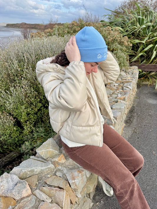 Person wearing a blue beanie and beige jacket sitting on a stone wall outdoors.