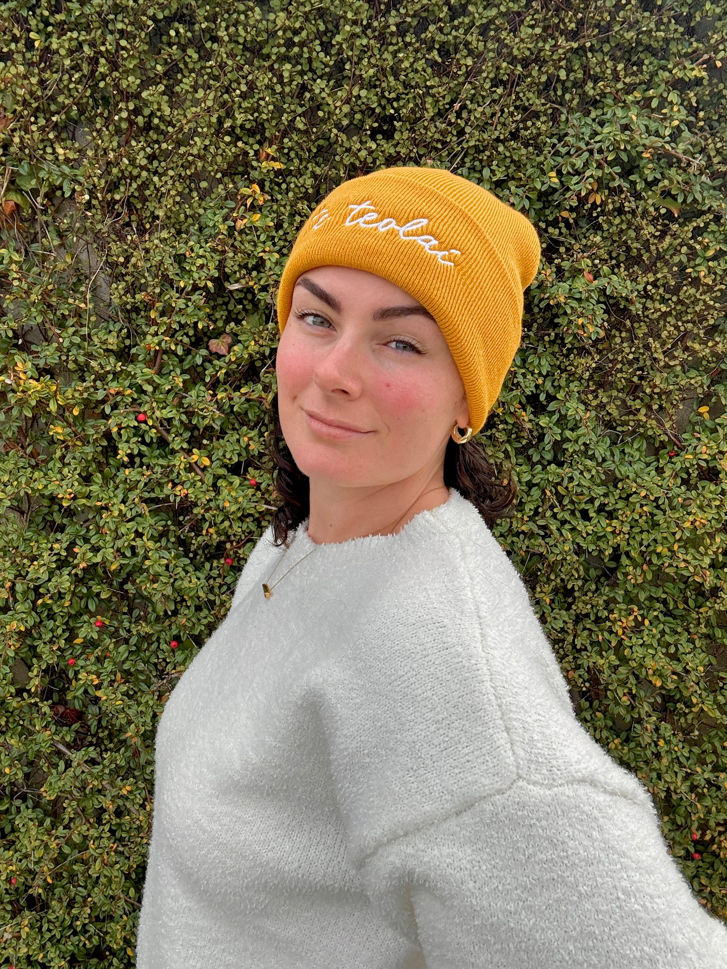 Person wearing a yellow beanie with 'te teolaí' branding against a green bush background