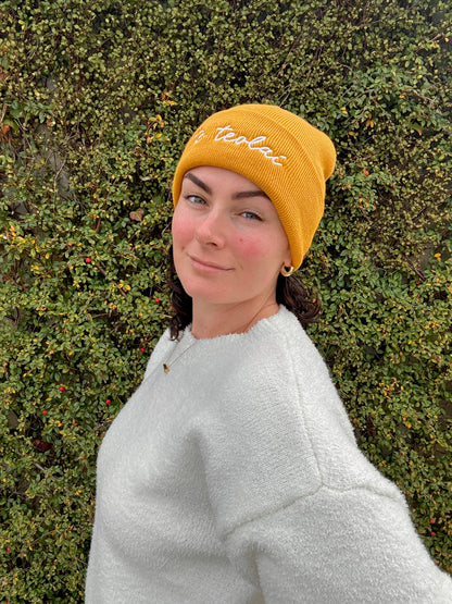 Person wearing a yellow beanie with 'te teolaí' branding against a green bush background